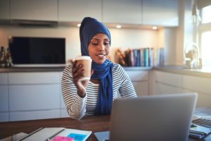 Young Arabic female entrepreneur wearing a hijab working on a laptop and drinking coffee while sitting at a table in her kitchen